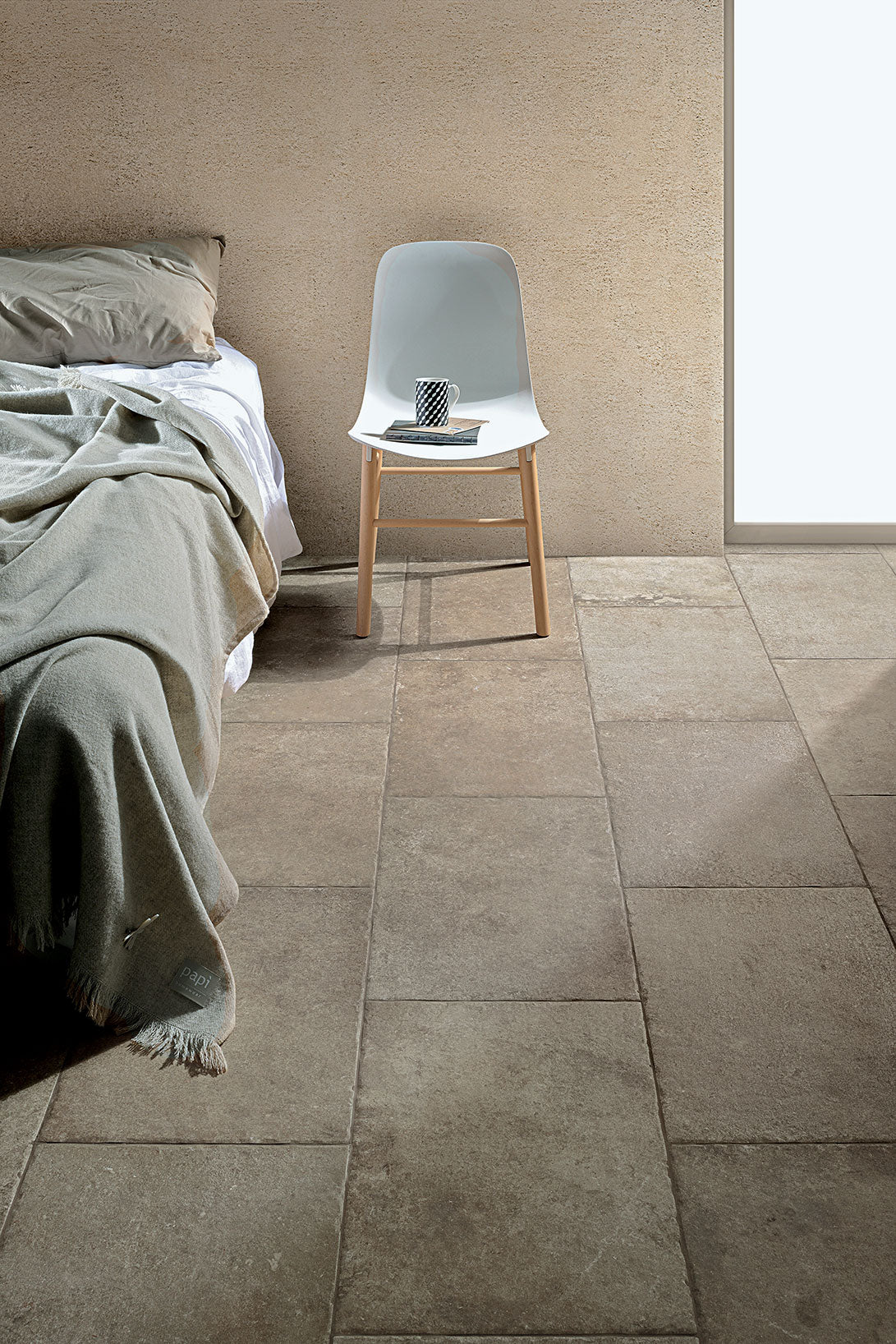 Lore taupe porcelain tile featured in a stylish bedroom setting with a cozy blanket and modern chair.